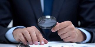 Man holding magnifying glass, examining document on clipboard.