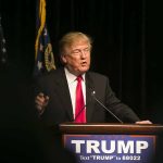 Man speaking at podium with "TRUMP" sign.