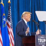 Person speaking at podium with flags in background.