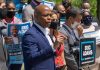 Man speaking at campaign event with supporters behind him.