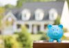 A blue piggy bank in front of a blurred house and greenery