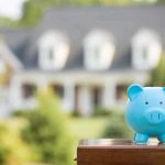 A blue piggy bank in front of a blurred house and greenery