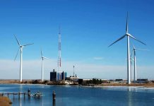 Wind turbines and a communication tower near a body of water under a clear blue sky