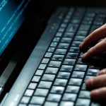 Close-up of hands typing on a laptop keyboard with code displayed on the screen