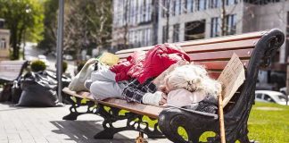 Person sleeping on park bench with cardboard sign.