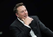 Musk’s $1 Trillion Payday Sparks Uprising – He Wants More! Man in suit smiling, resting chin on hand.