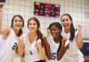 Four female volleyball players celebrating in a gymnasium