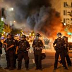 Man Moons Protesters: Humiliating Window Surprise Police officers in riot gear near burning car.