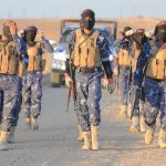 Camouflaged soldiers wearing masks and vests walk in desert.