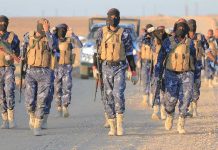 Camouflaged soldiers wearing masks and vests walk in desert.