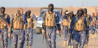Camouflaged soldiers wearing masks and vests walk in desert.