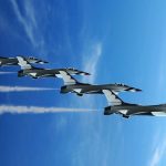 Four military jets flying in formation against a blue sky, leaving smoke trails