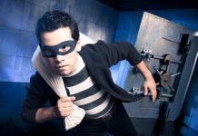 A masked thief in a striped shirt holding a bag while emerging from a bank vault
