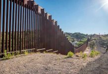 Tall metal border wall with rural landscape.