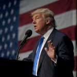 man in suit speaking at a podium with a US flag backdrop