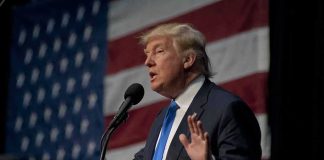 man in suit speaking at a podium with a US flag backdrop