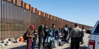 Group of people standing by a border fence with officers and vehicles