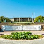 Government building behind landscaped gardens and a stone relief wall